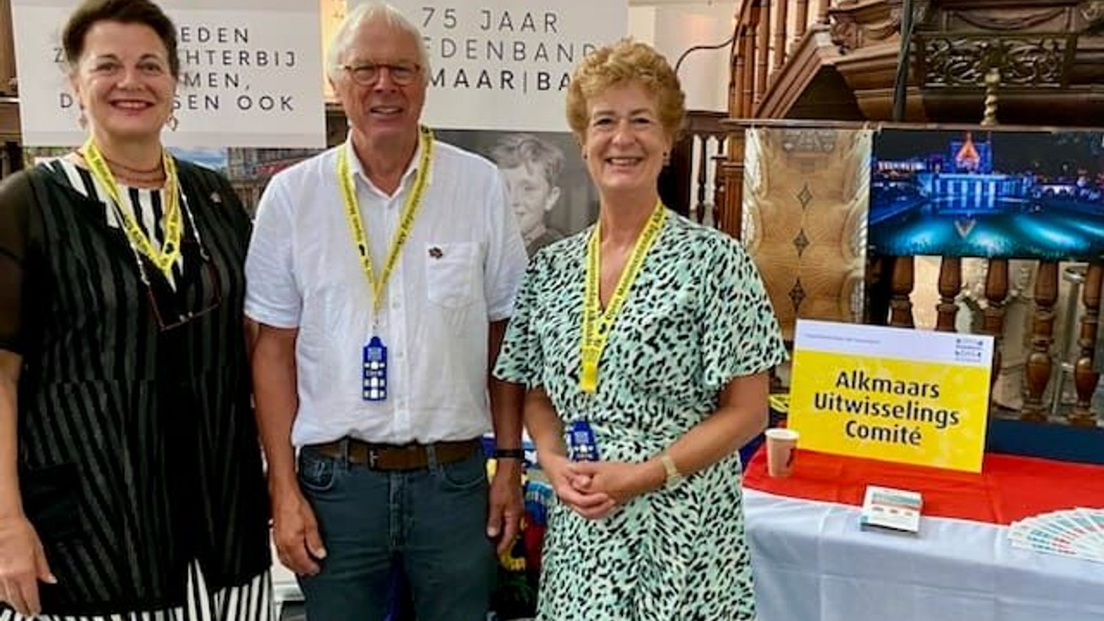 Drie personen in gele naambandje staan achter rode tafel op binnenevenement met tentoonstellingsborden op achtergrond