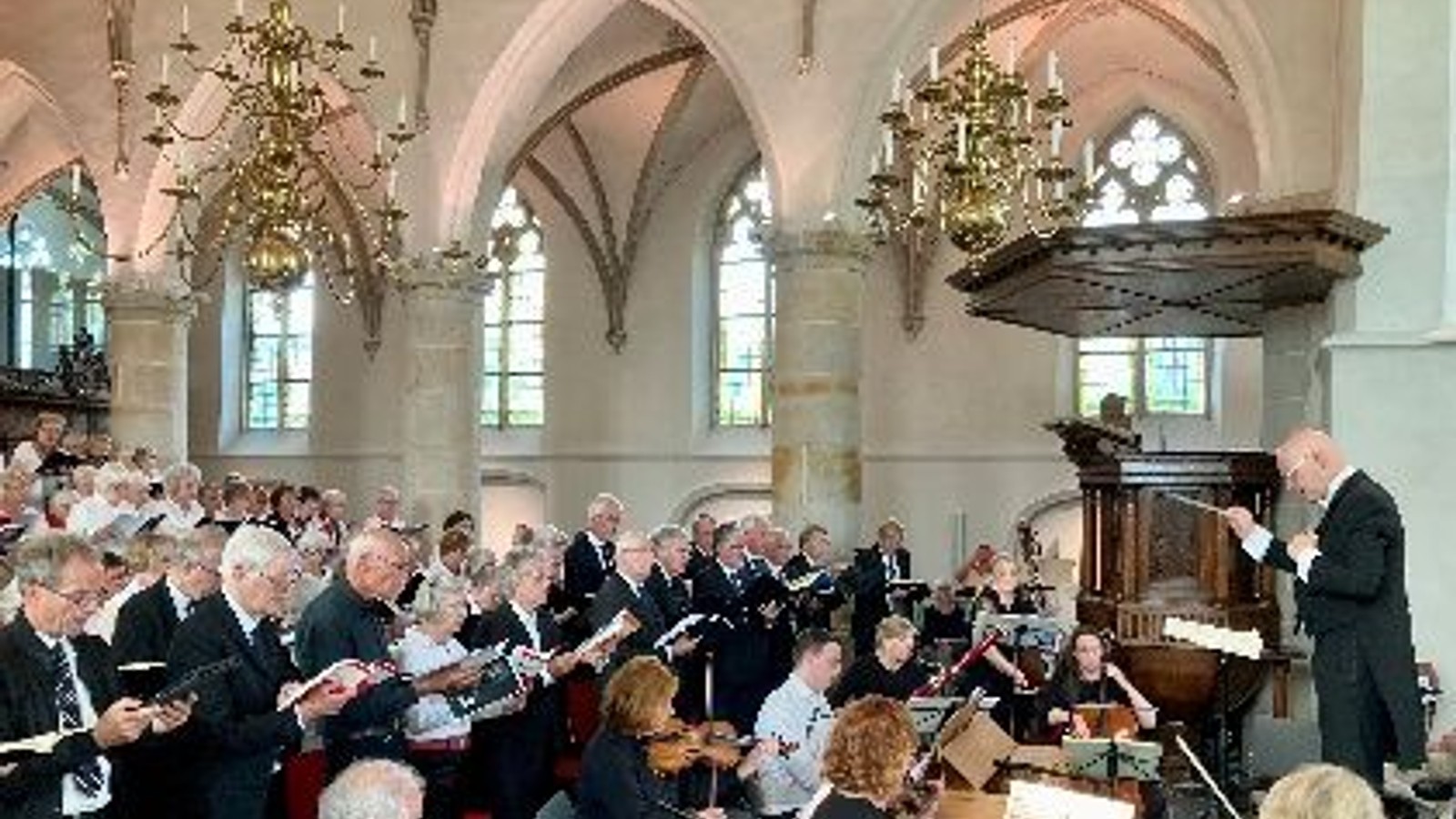 Grote groep zangeres met bladmuziek in kerkinterieur met gewelven, kroonluchters en dirigent.