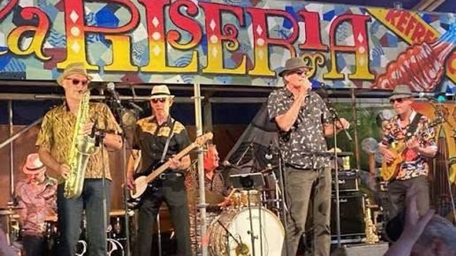 Muziekband met vijf leden speelt op podium onder kleurrijk bord met 'Allski' tekst, enkele leden dragen gekleurde kleding en hoeden