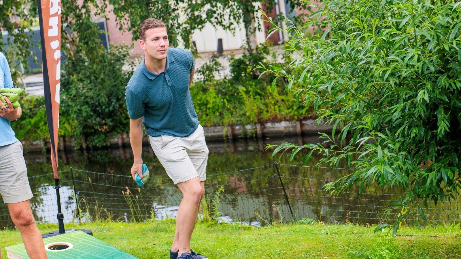 Man in blauw shirt gooit cornhole zak naar doelkuil op gras aan waterkant met begroeiing