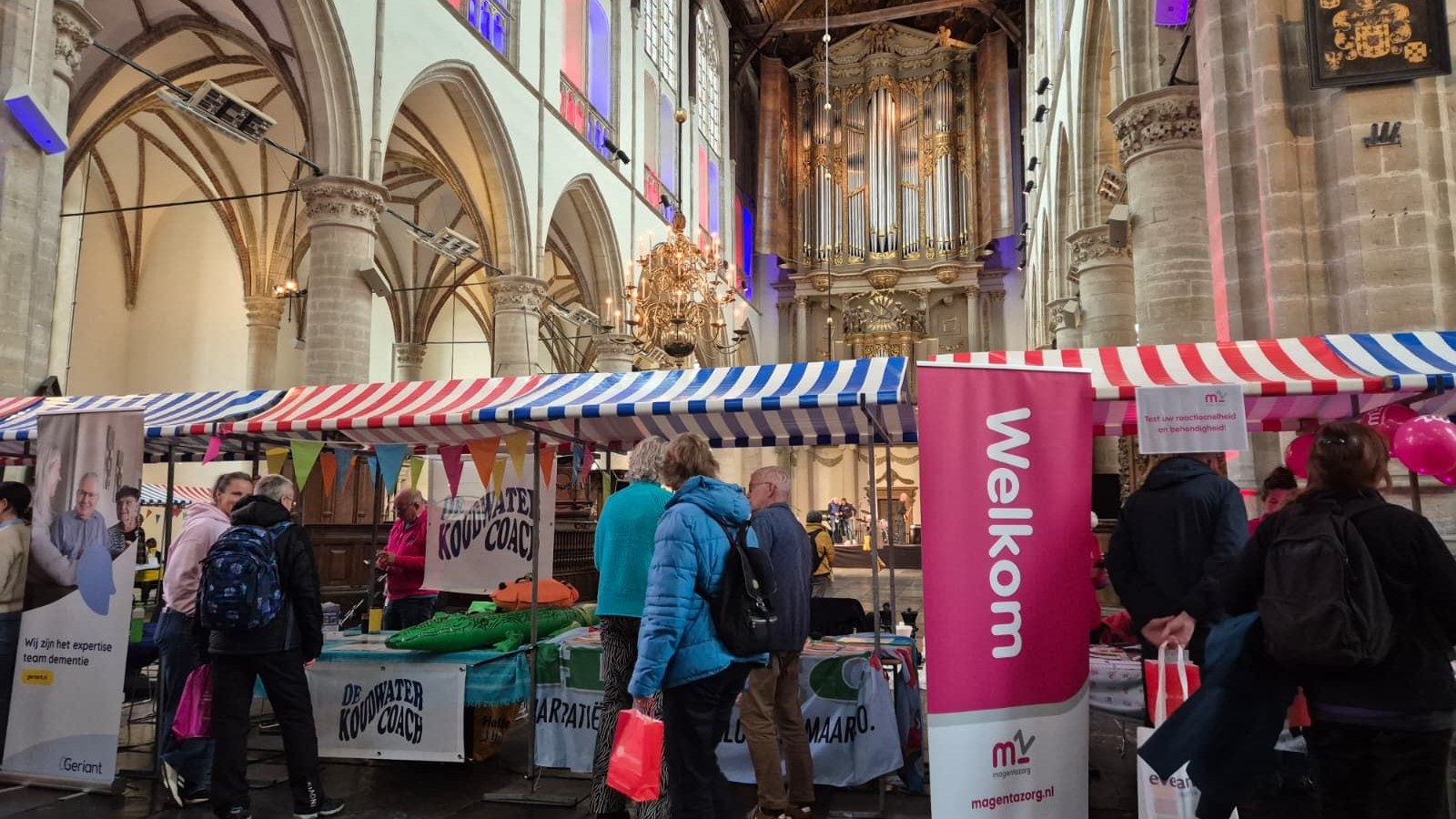 Binnenkant kerk met bezoekers bij marktstandjes onder witte en blauwe gestreepte parasols, orgel op achtergrond.