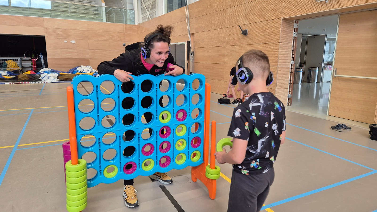 Twee kinderen spelen vier-op-een-rij spel in een sporthal, beide dragen koptelefoons, omringende speeltoestellen zichtbaar.