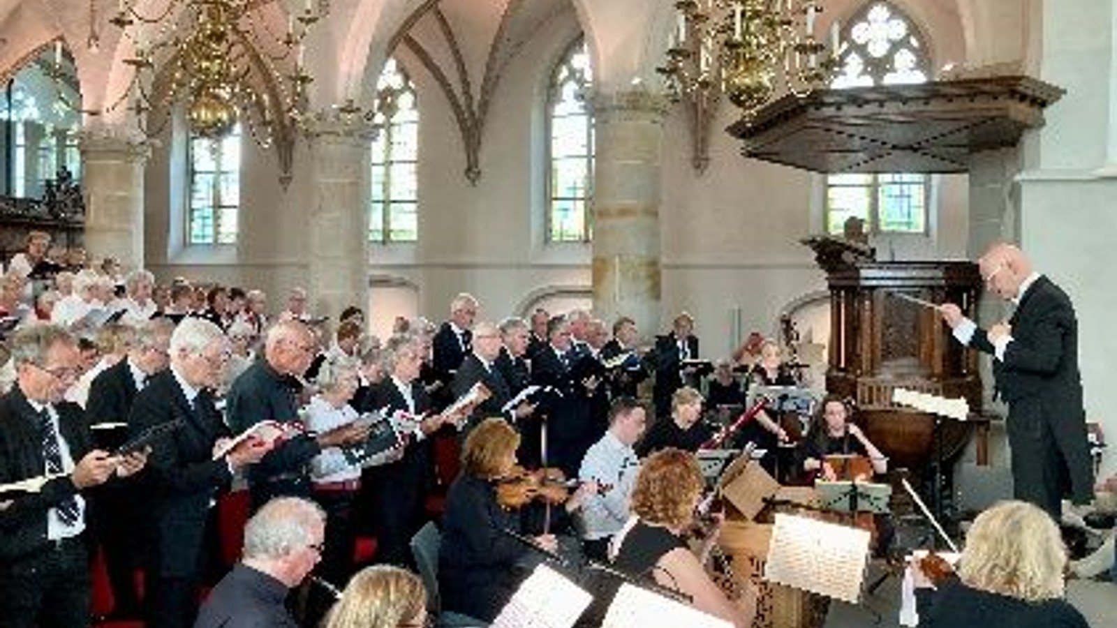 Kerkinterieur vol zangers in donkere kleding met partituren, dirigent leidt aan podium, orgel en ramen zichtbaar