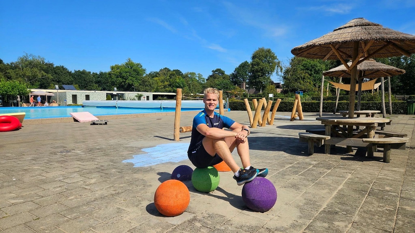 Persoon zittend op gekleurde bal op terras met zwembad, speelelementen en parasol op achtergrond