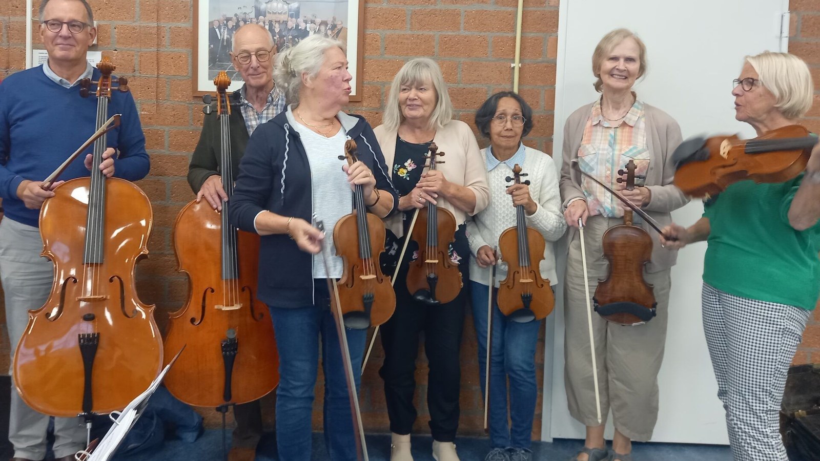 Zes mensen met strijkinstrumenten staan naast elkaar in een bakstenen ruimte