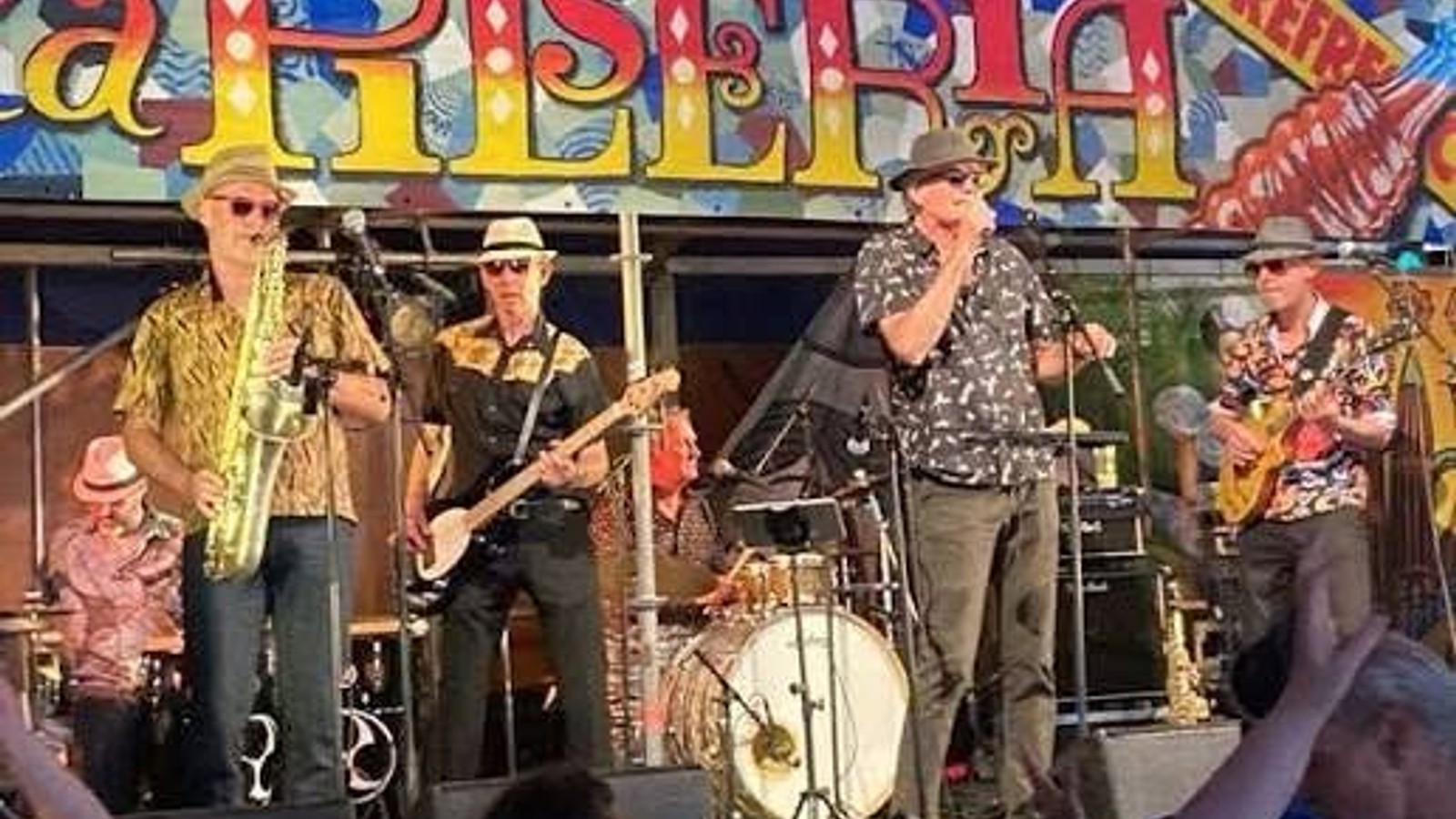 Muziekband met vijf leden speelt op podium onder kleurrijk bord met 'Allski' tekst, enkele leden dragen gekleurde kleding en hoeden