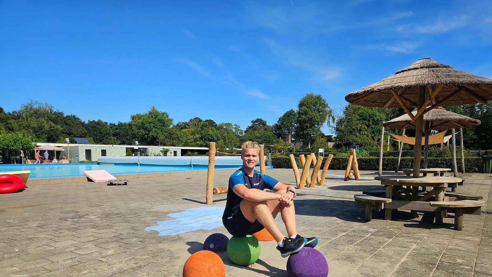 Buitenbad terras met zwembad, houten speelelementen, gekleurde ballen, parasol en omringende bomen op zonnige dag