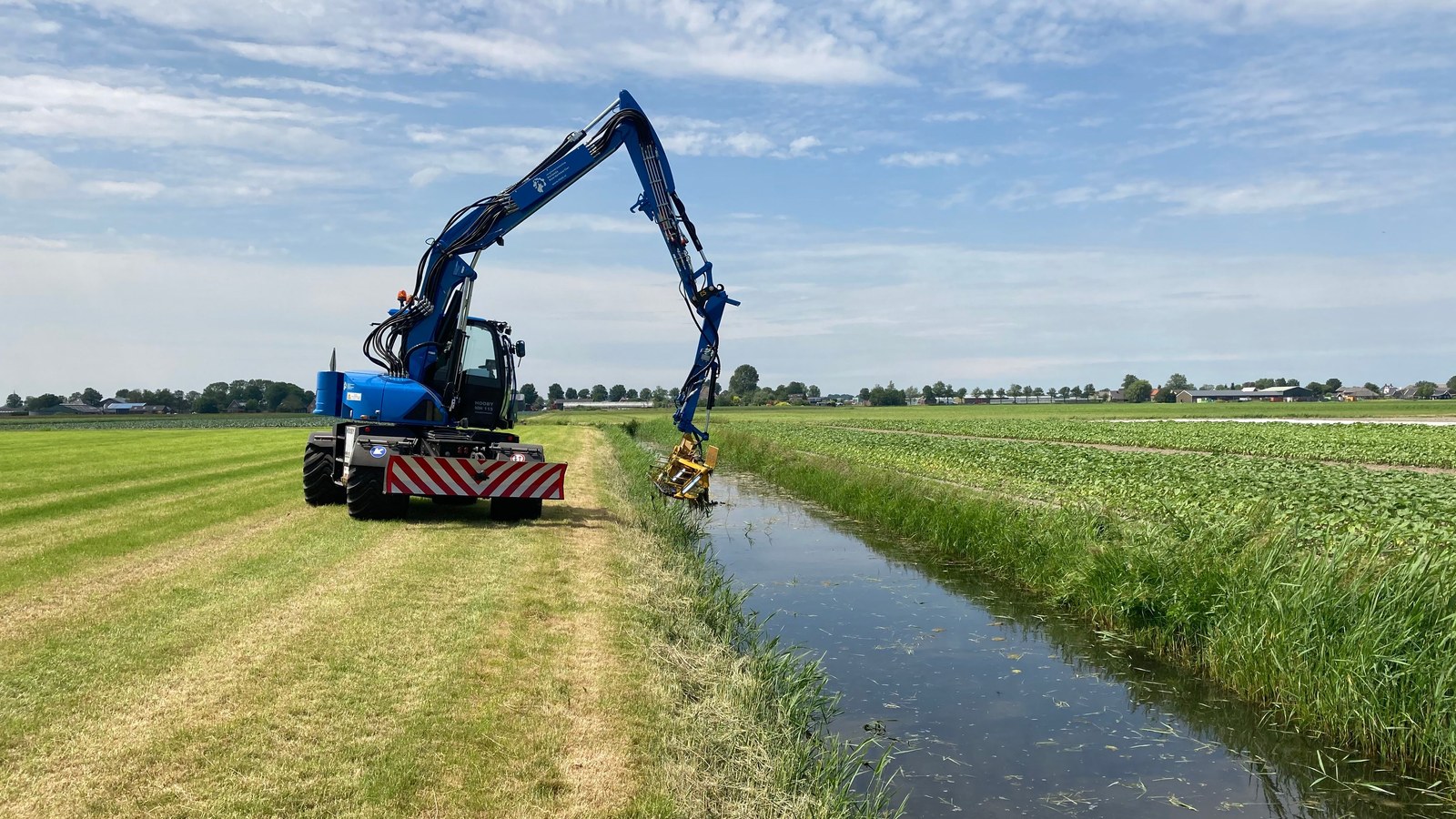 Blauwe maaimachine met lange arm aan slootkant in landbouwlandschap tussen groene akkers.