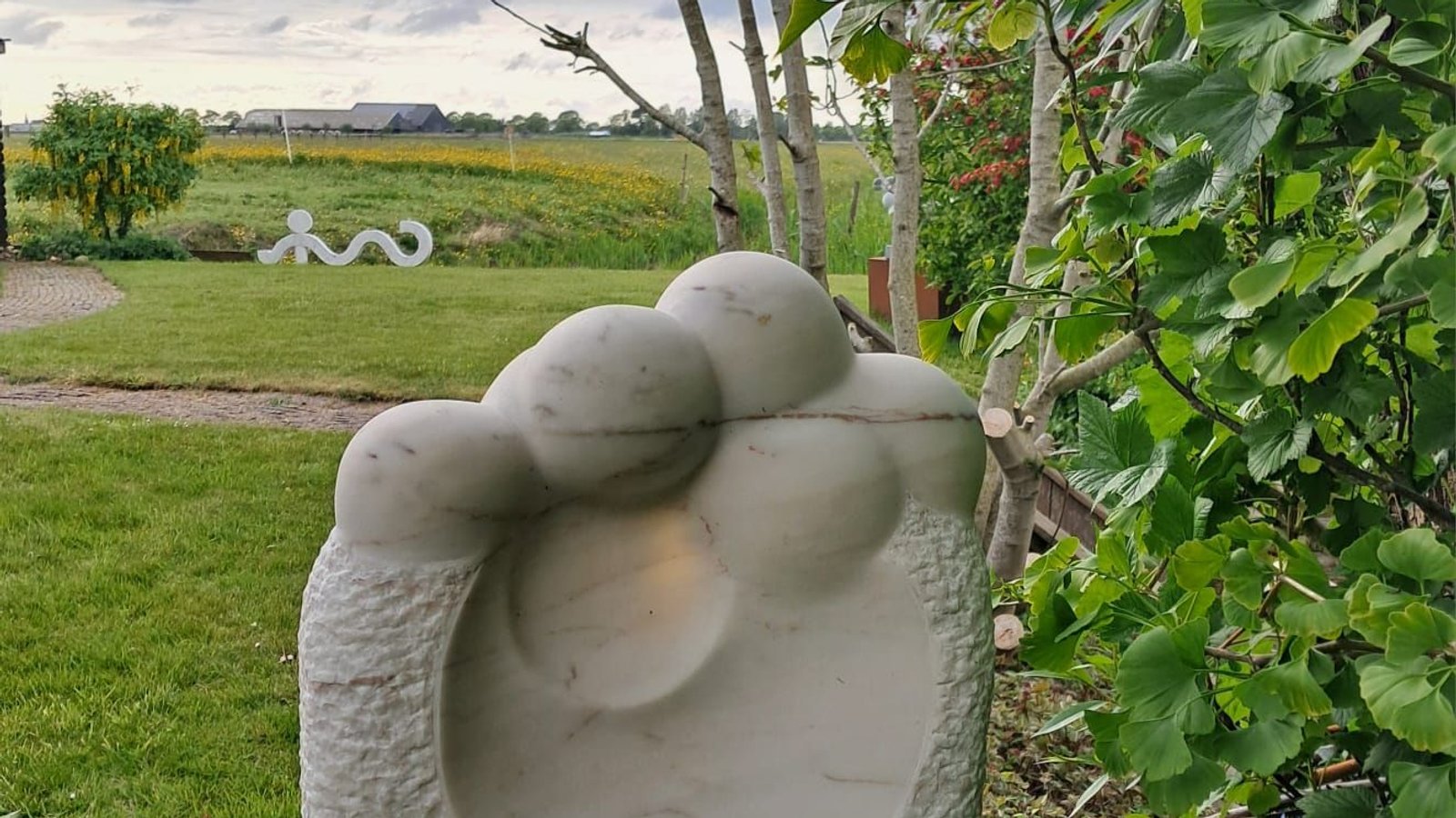 Witte stenen sculptuur van abstracte vorm in tuin naast boom en groene bladeren.