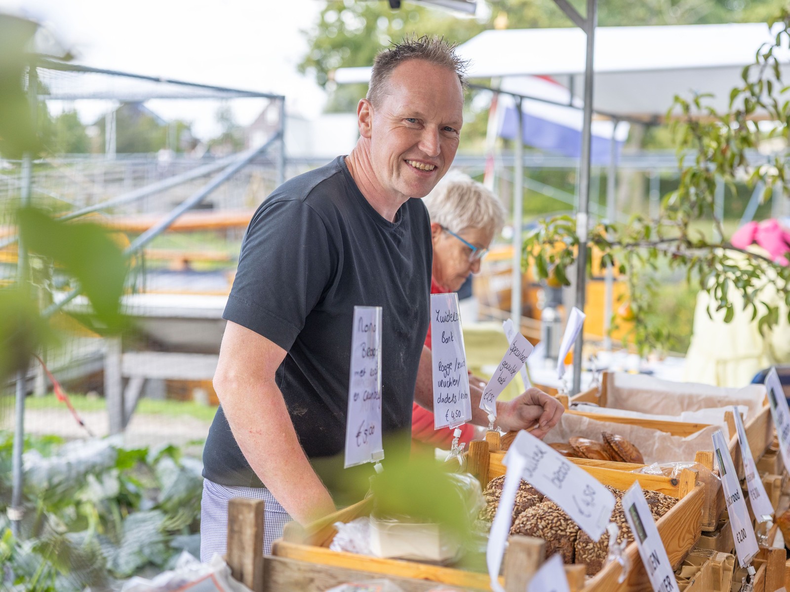 Man in zwart shirt staat achter marktstand met houten bakken vol producten en prijskaartjes, in kas-omgeving van museumterrein