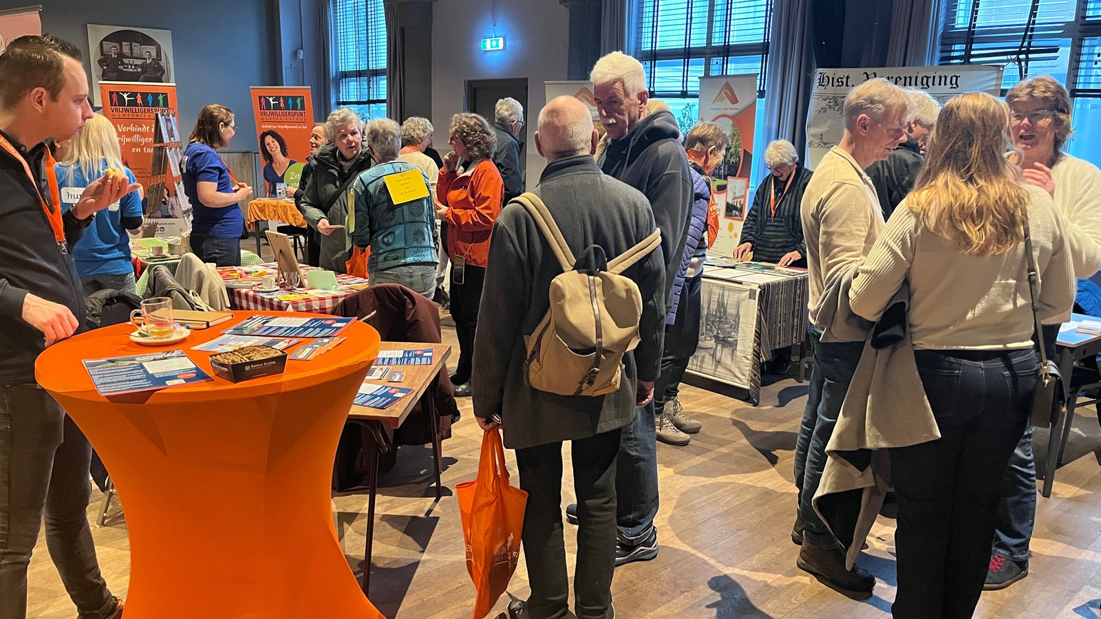 Groep bezoekers en deelnemers bij oranje tafels en informatiestandaard op een vrijwilligersmarkt in een zalenruimte