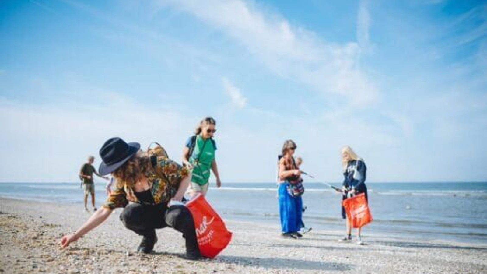 Meerdere personen verzamelen afval op een strand. Een persoon in het midden bukt zich met een rode zak, terwijl anderen in de buurt staan