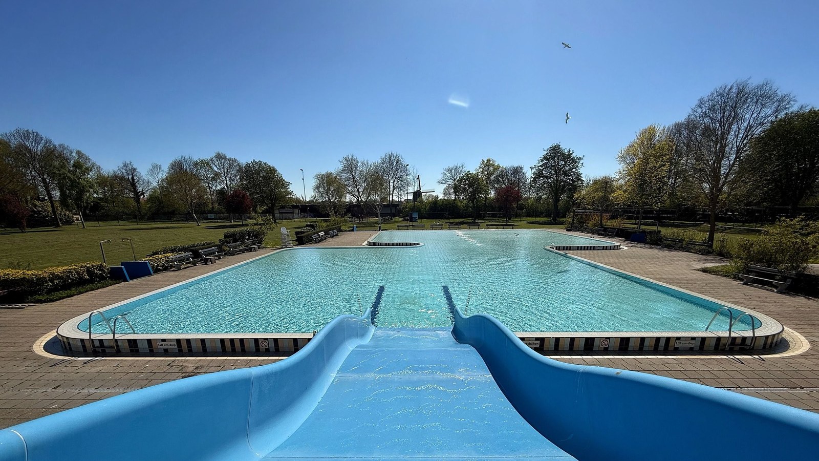Rechthoekig buitenbad met turquoise water en twee blauwe glijbanen vanaf het dek gezien, omgeven door groene grasvelden en bomen.