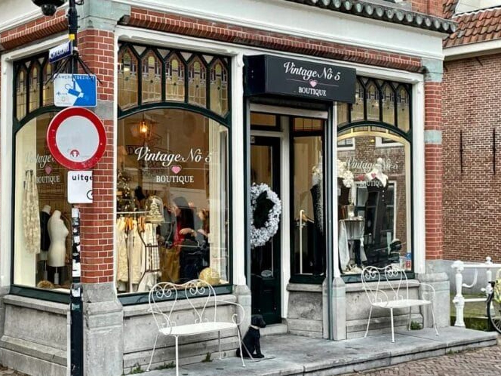 Vintage kledingboutique aan een bakstenen straatgevel met uitstalramen, mannequin in etalage en witte tuinstoelen buiten.