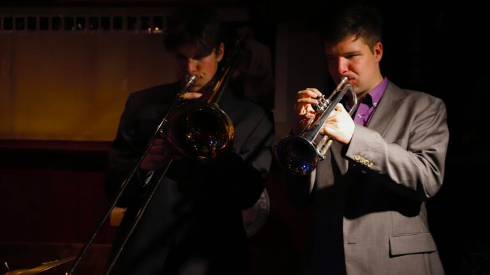 Twee jazzmusici spelen samen op een podium: links een musicus met trombone in zwart pak, rechts een musicus met trompet in grijs pak met