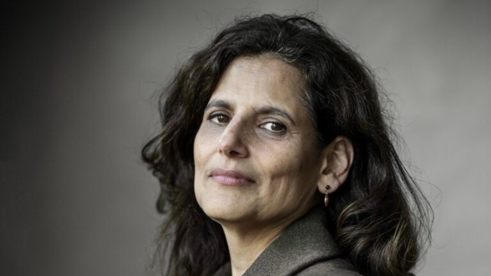 Portretfoto vrouw met donker golvend haar en grijze blouse tegen grijze achtergrond, kijkend naar camera