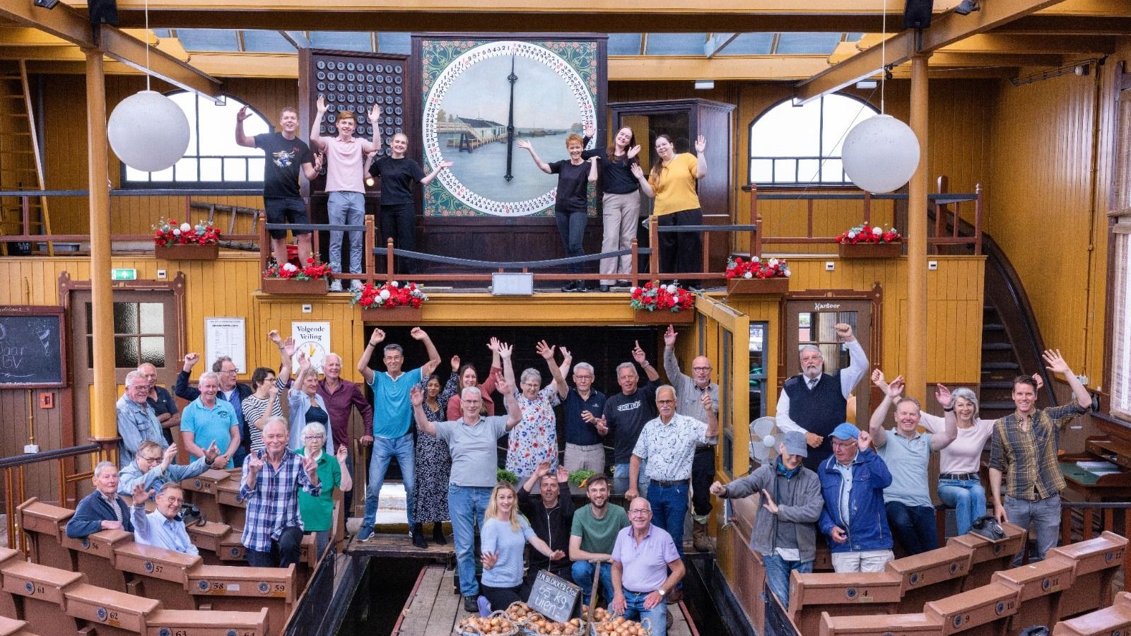 Groep mensen zwaait naar camera in houten interieur met grote ronde klok en bloemenversiering