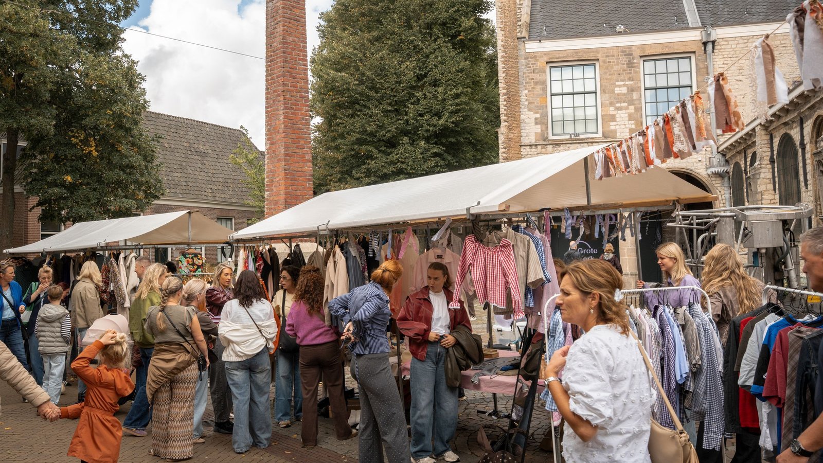 Overdekte markt met veel bezoekers tussen kraampjes met kleding aan rekken onder witte tenten, bakstenen gebouwen op achtergrond