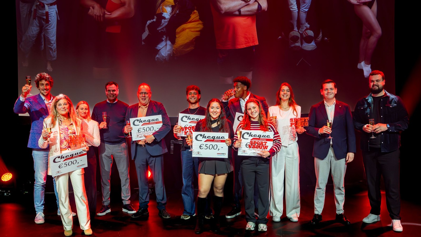 Groep personen op podium onder rood licht, enkele houden cheques van 500 euro omhoog