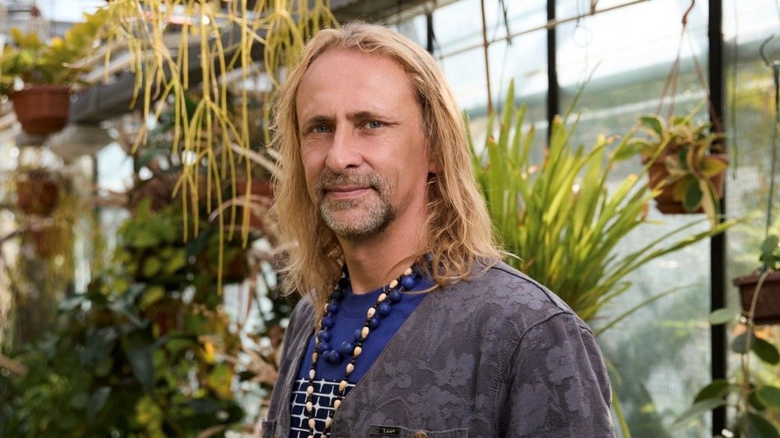 Man met lang blond haar in blauw shirt staat tussen hangende en potplanten in kas