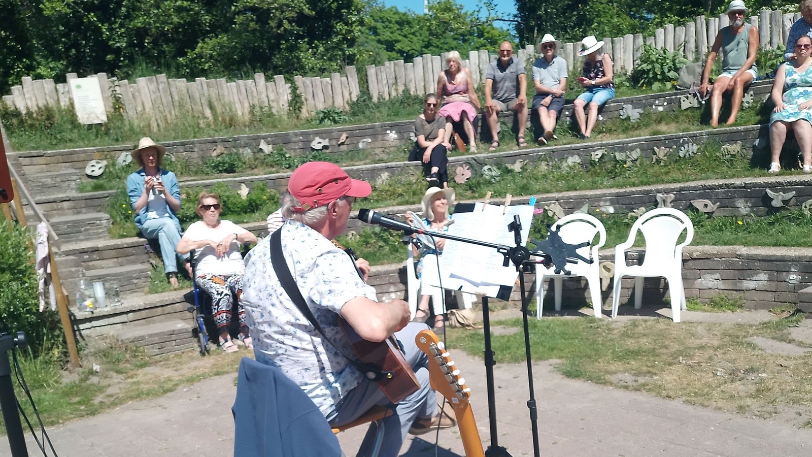 Persoon met rode pet speelt gitaar op een podium in een park. Zittend publiek luistert naar het optreden op houten zitbanken.