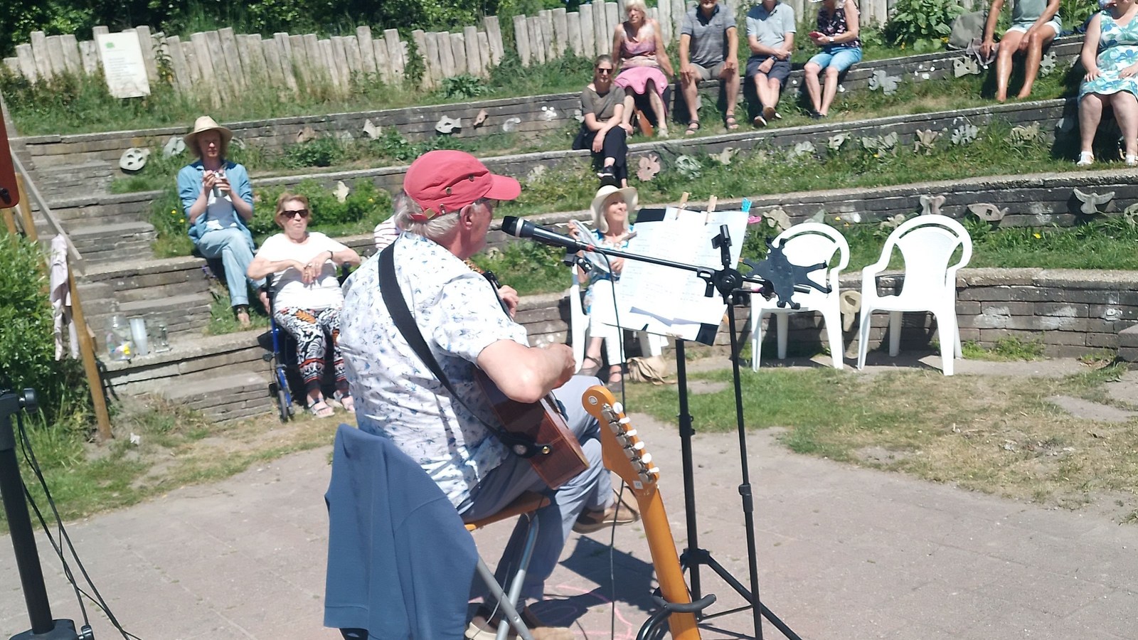 Persoon met rode pet speelt gitaar op een podium in een park. Zittend publiek luistert naar het optreden op houten zitbanken.