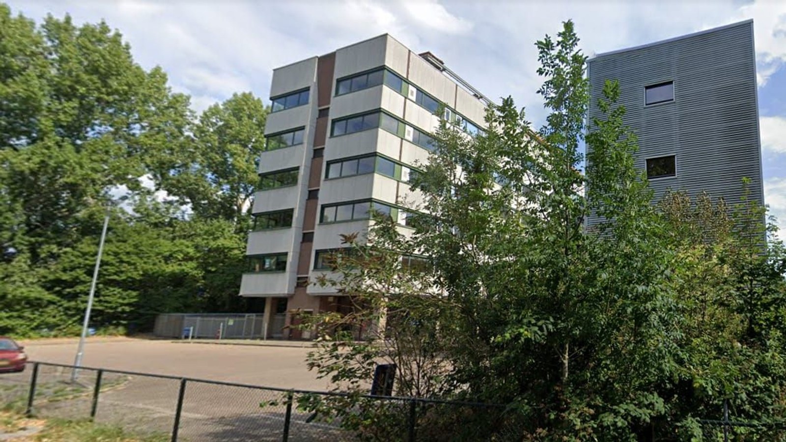 Voorkant van een modern appartementengebouw met meerdere verdiepingen, grijze en bruine gevel, grote ramen, omgeven door groene bomen en