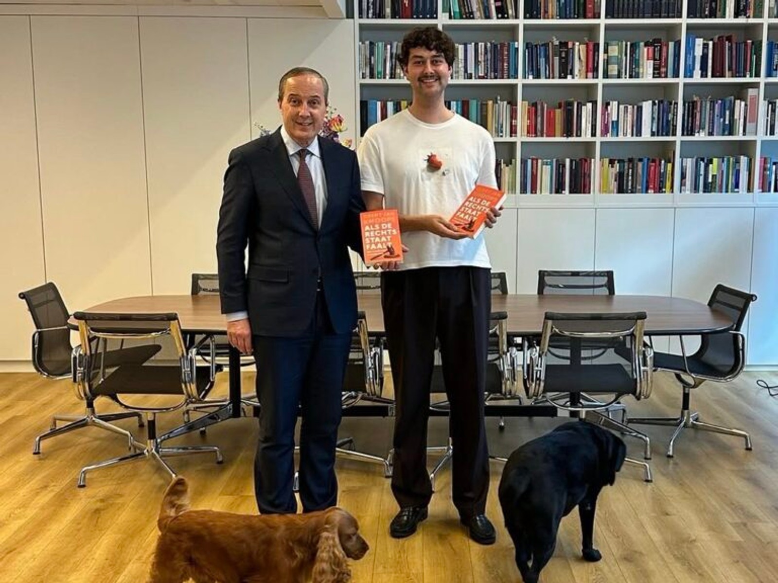 Man in donker pak en man in wit shirt houden rode boeken vast in kantoor met boekenkasten en twee honden