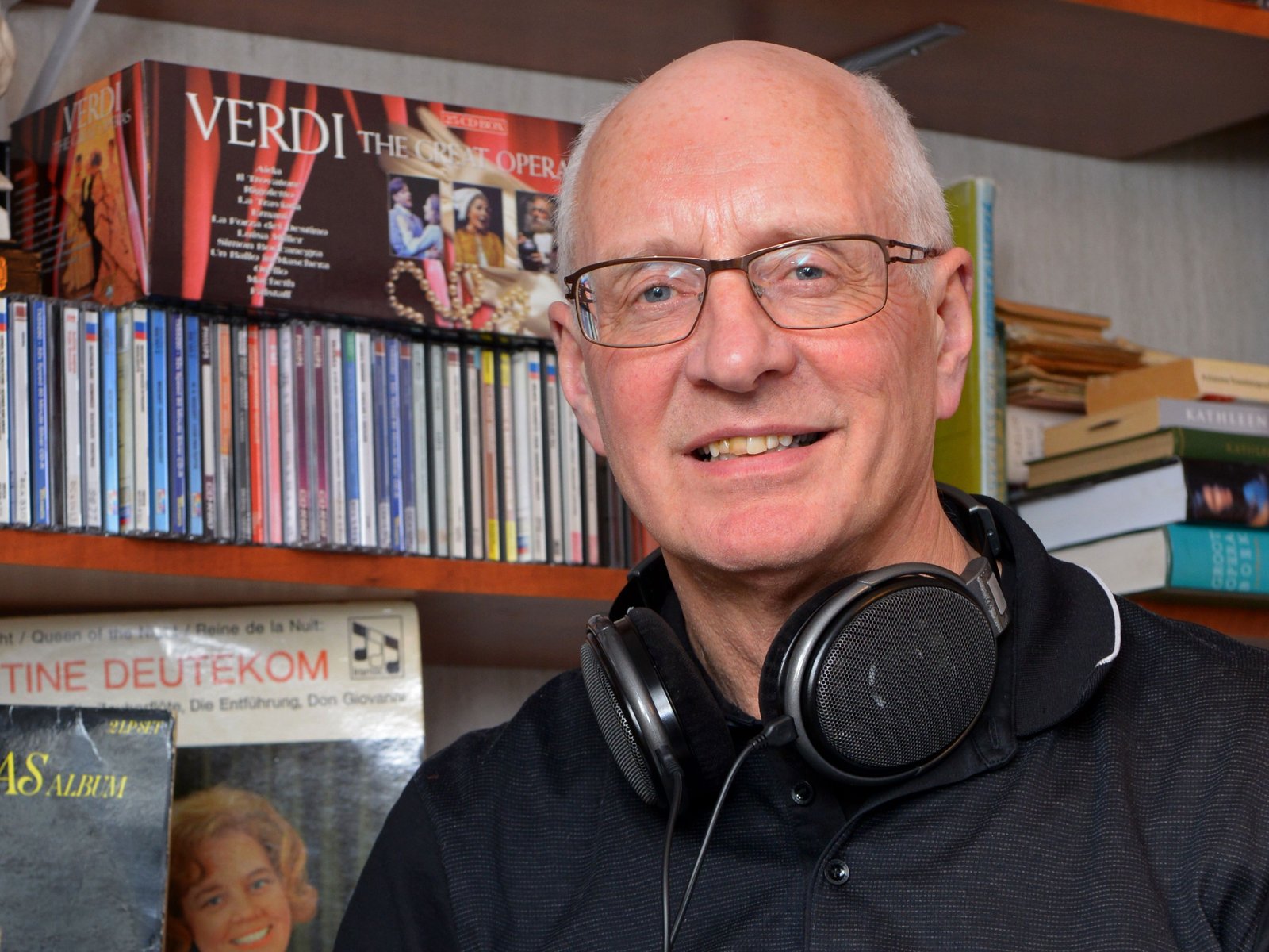 Persoon met bril en koptelefoon zit voor boekenkast met cd's en dvd's, Verdi opera poster op achtergrond