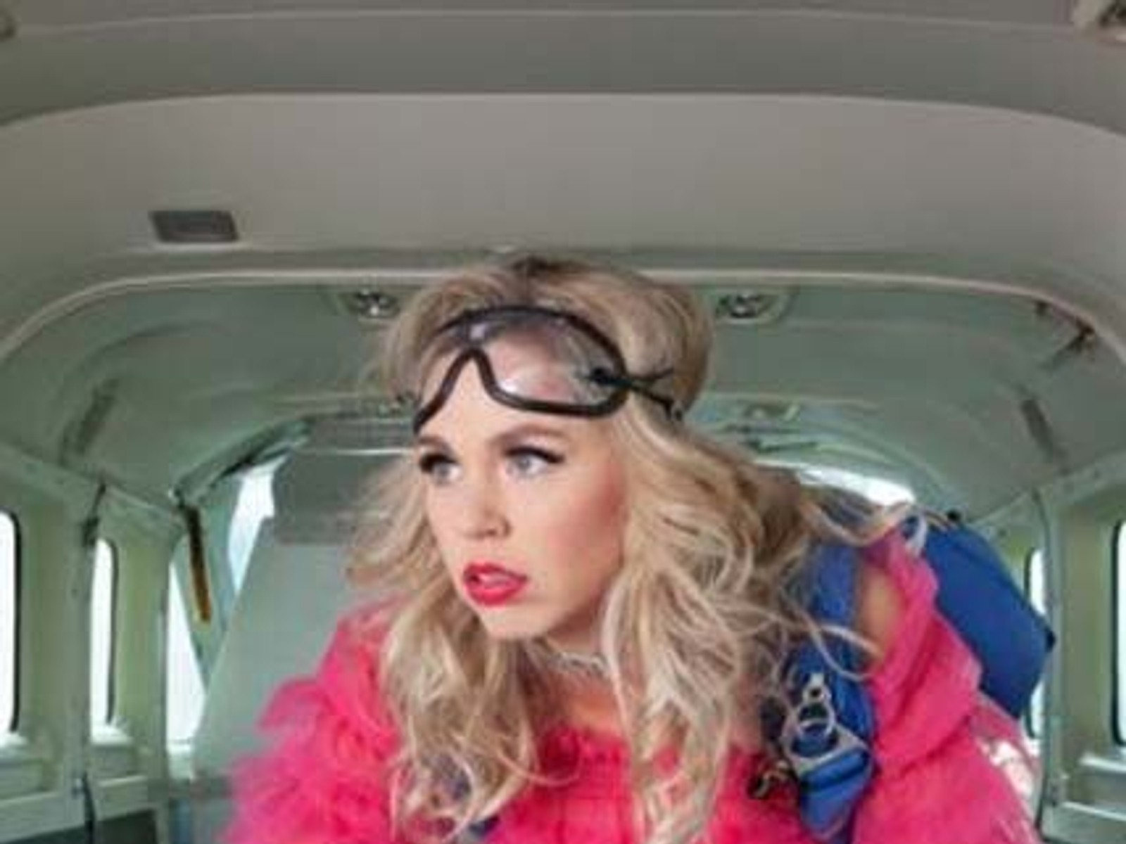 Persoon met blonde krullen en bril op het hoofd zit in een vintage auto met groen interieur, draagt een fel roze jas