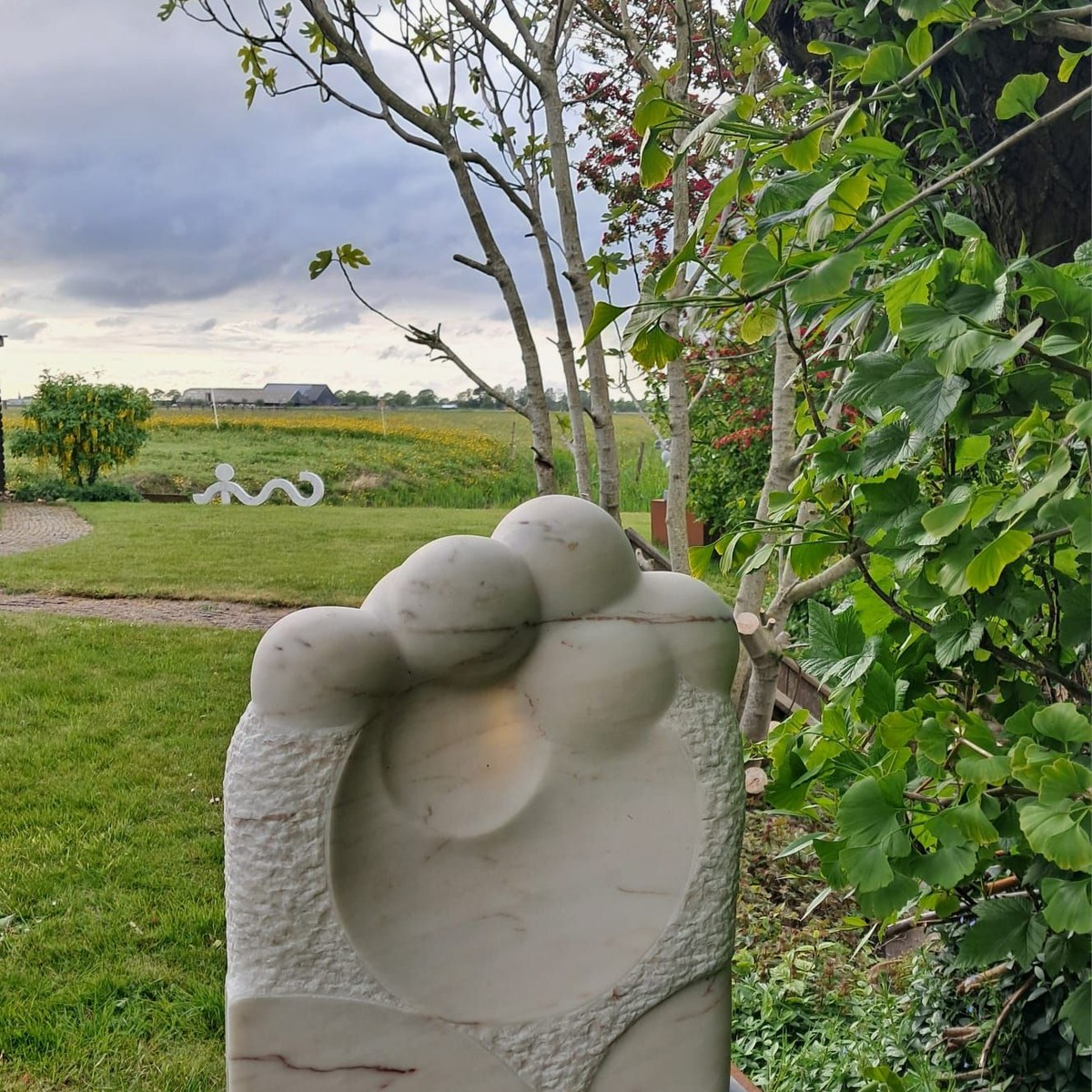 Witte stenen sculptuur van abstracte vorm in tuin naast boom en groene bladeren.