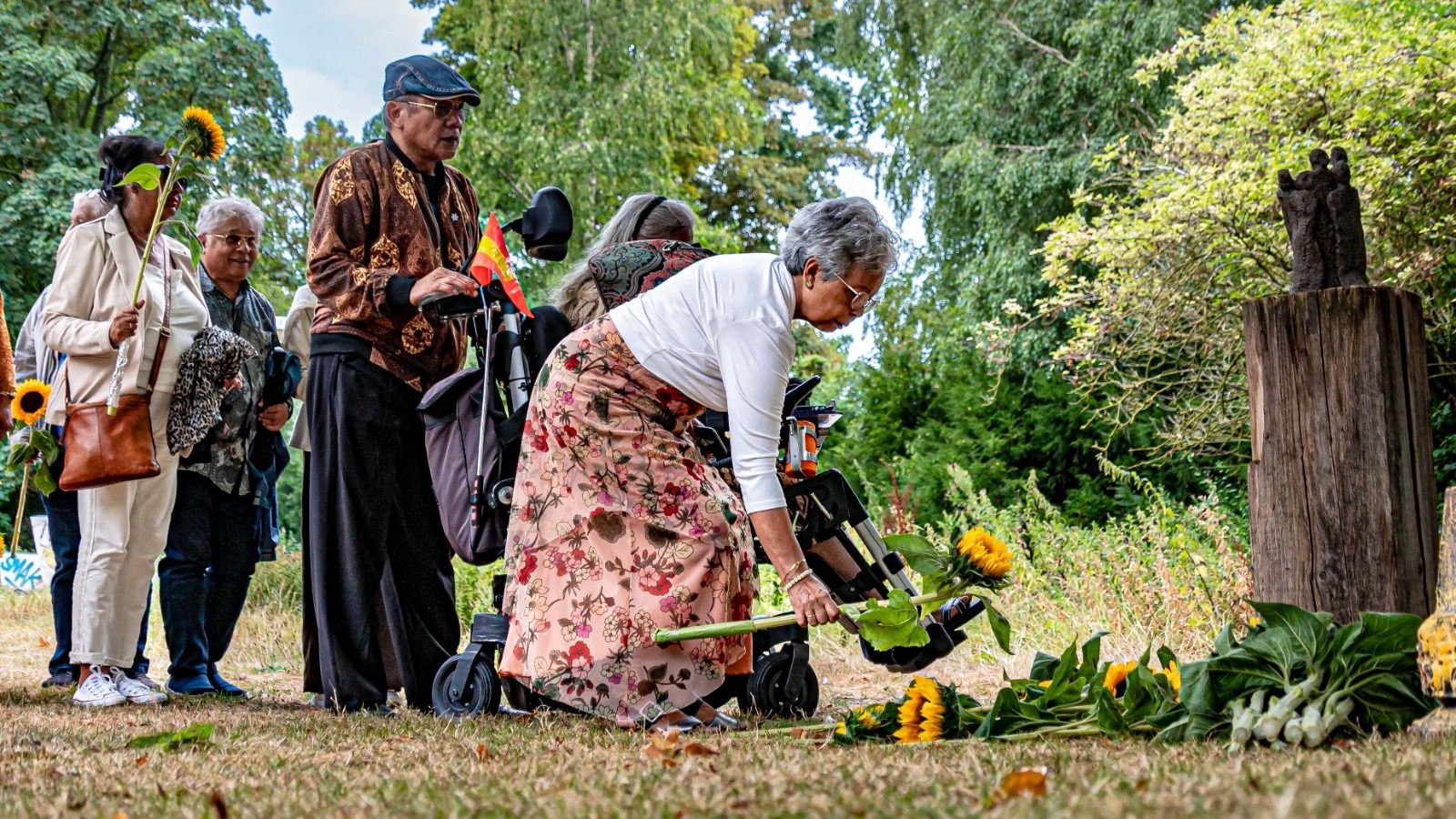 Groep mensen bij houten monument in park, vrouw plaatst bloemen op gras tussen zonnebloemen, opgaande bomen op achtergrond