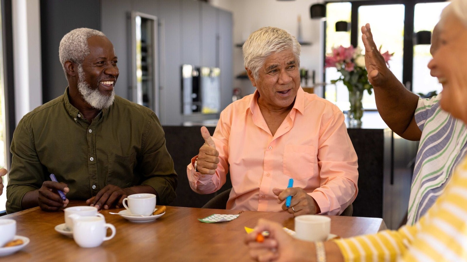 Drie volwassenen zitten aan een tafel met kopjes koffie en hebben een gesprek in een interieur met bloemen op de achtergrond.