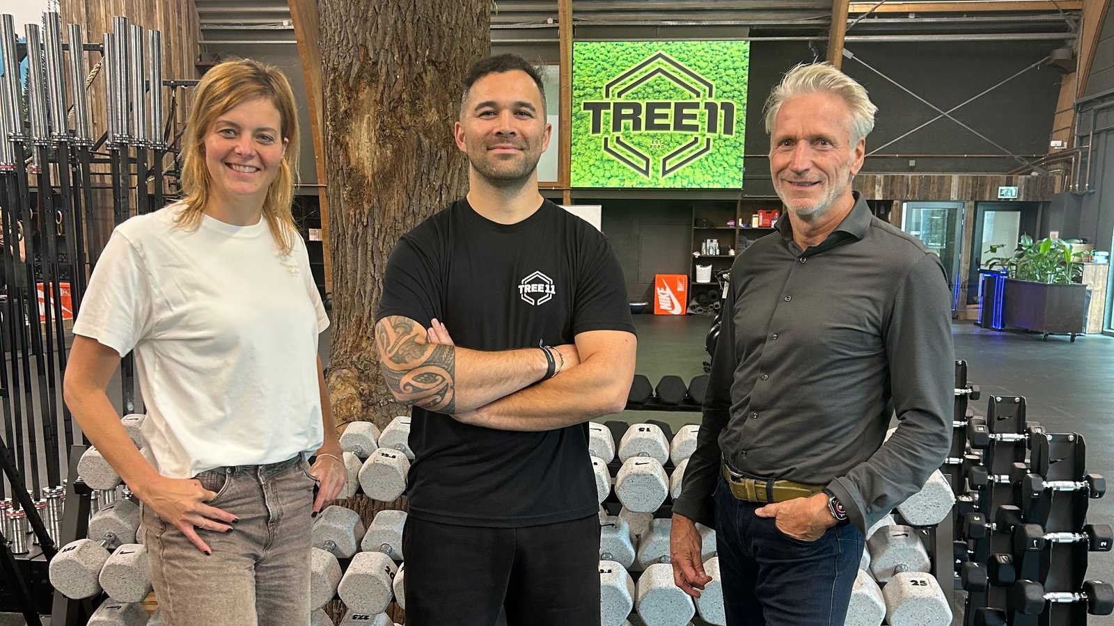 Drie personen poseren in trainingsfaciliteit tussen halters, TREE11-logo zichtbaar op muur, zijkant met trainingsapparatuur.