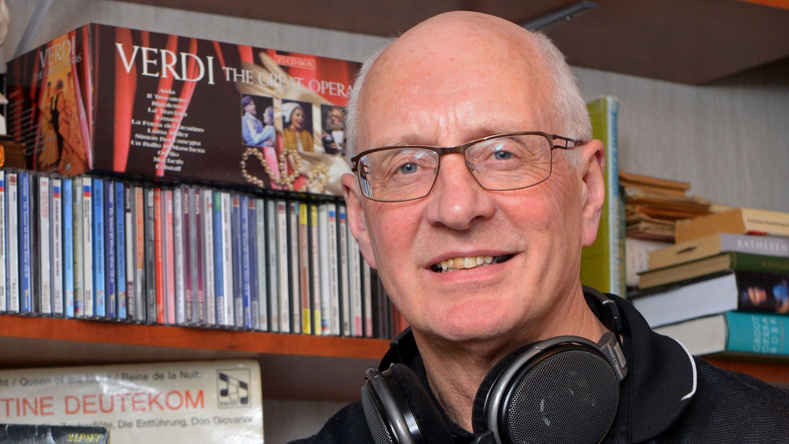 Persoon met bril en koptelefoon om nek zit voor boekenkast met cd's en dvd's, Verdi opera poster zichtbaar