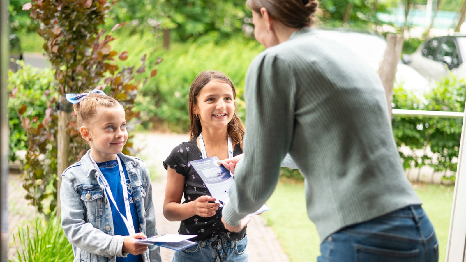 Twee kinderen met halsbanden geven lotjes aan een volwassene in een tuin, voor huizen en groen