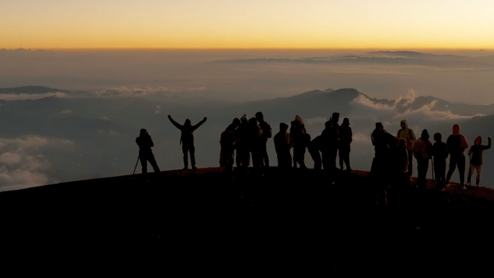 Silhouetten van ongeveer twintig personen staan op een bergkam tegen een zonsopganghemel met wolken.