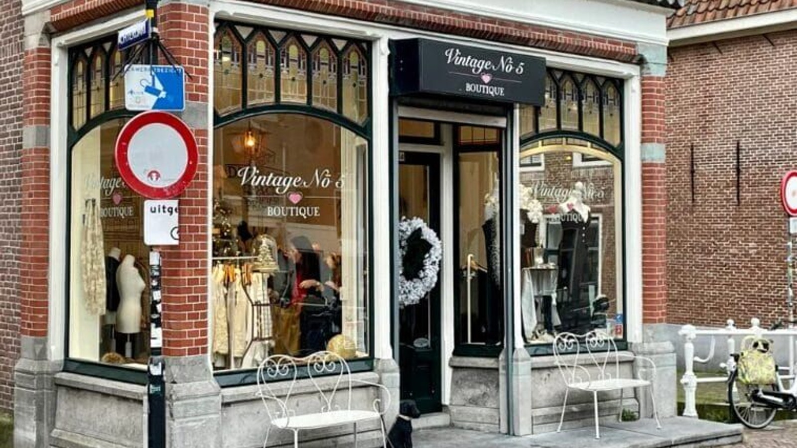 Vintage kledingboutique aan een bakstenen straatgevel met uitstalramen, mannequin in etalage en witte tuinstoelen buiten.