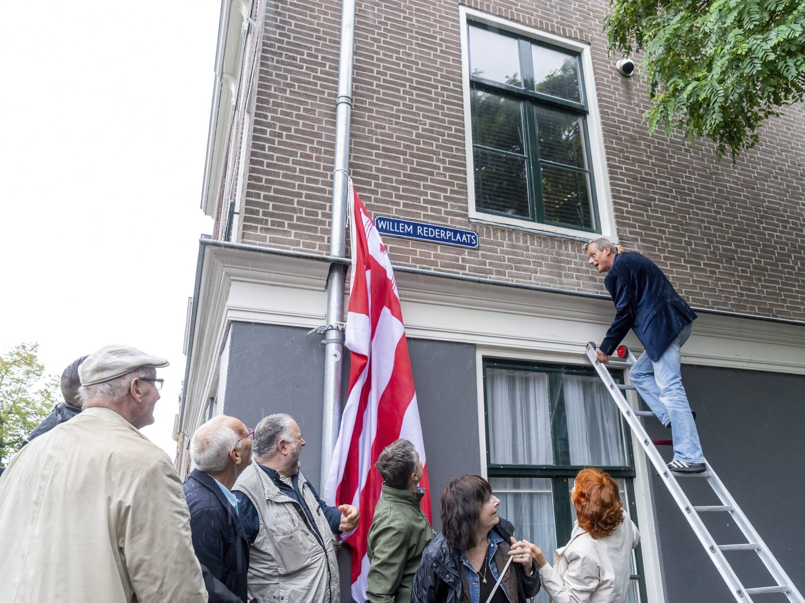 Man op ladder onthult straatnaambord Willem Rederplaats aan bakstenen gebouw met Nederlandse vlag en groep toeschouwers eronder.