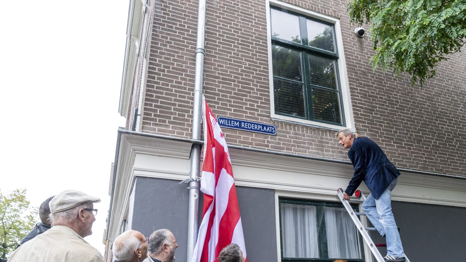 Man op ladder onthult straatnaambord Willem Rederplaats aan bakstenen gebouw met Nederlandse vlag, omringd door toeschouwers.
