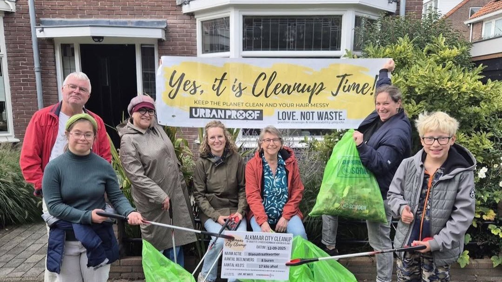 Groep van zes personen staat buiten voor een huis met een banner 'Yes it is Cleanup Time' en houdt opruimgereedschap vast.