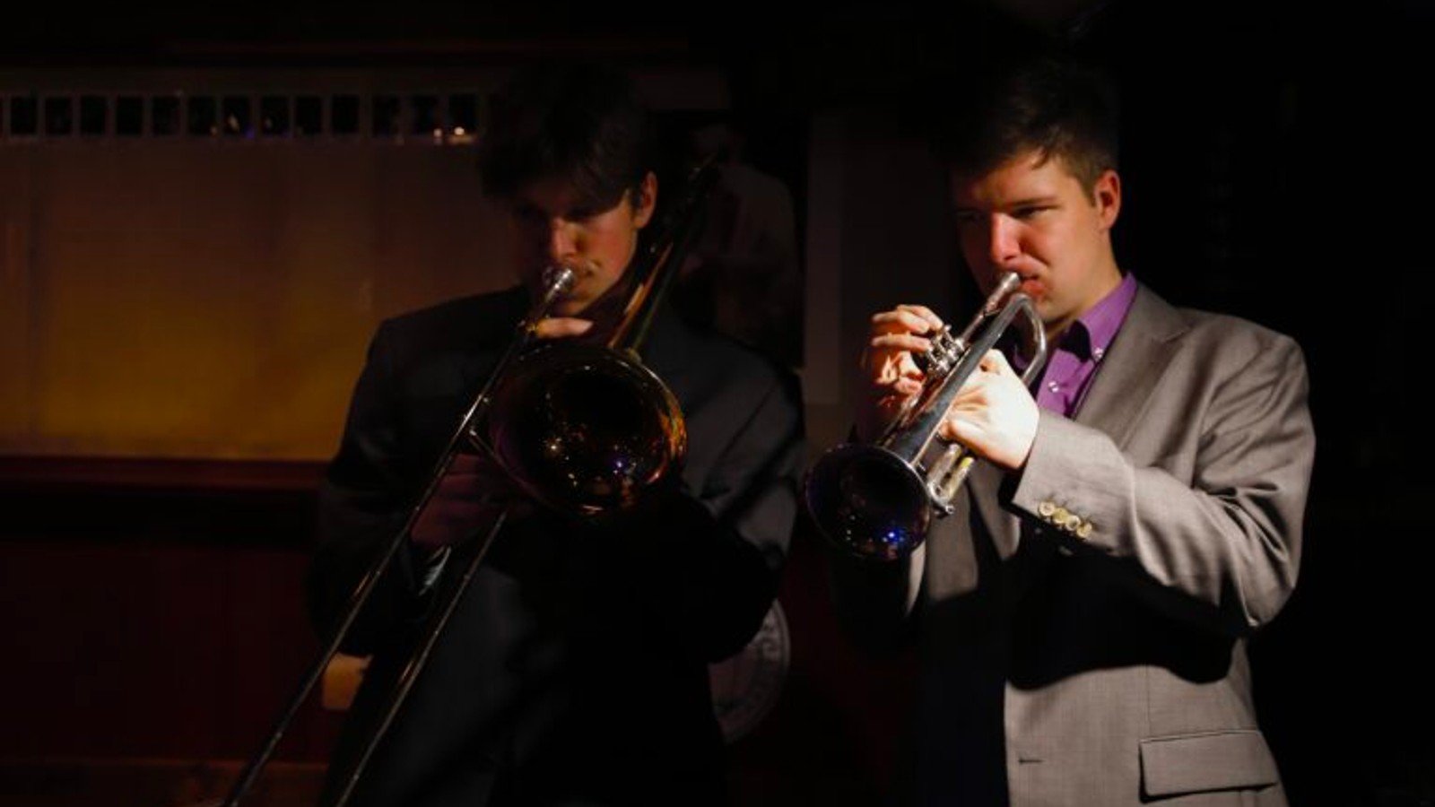 Twee muzikanten spelen op een podium, links een man met trombone in zwart pak, rechts een man met trompet in grijs pak met paars overhemd.