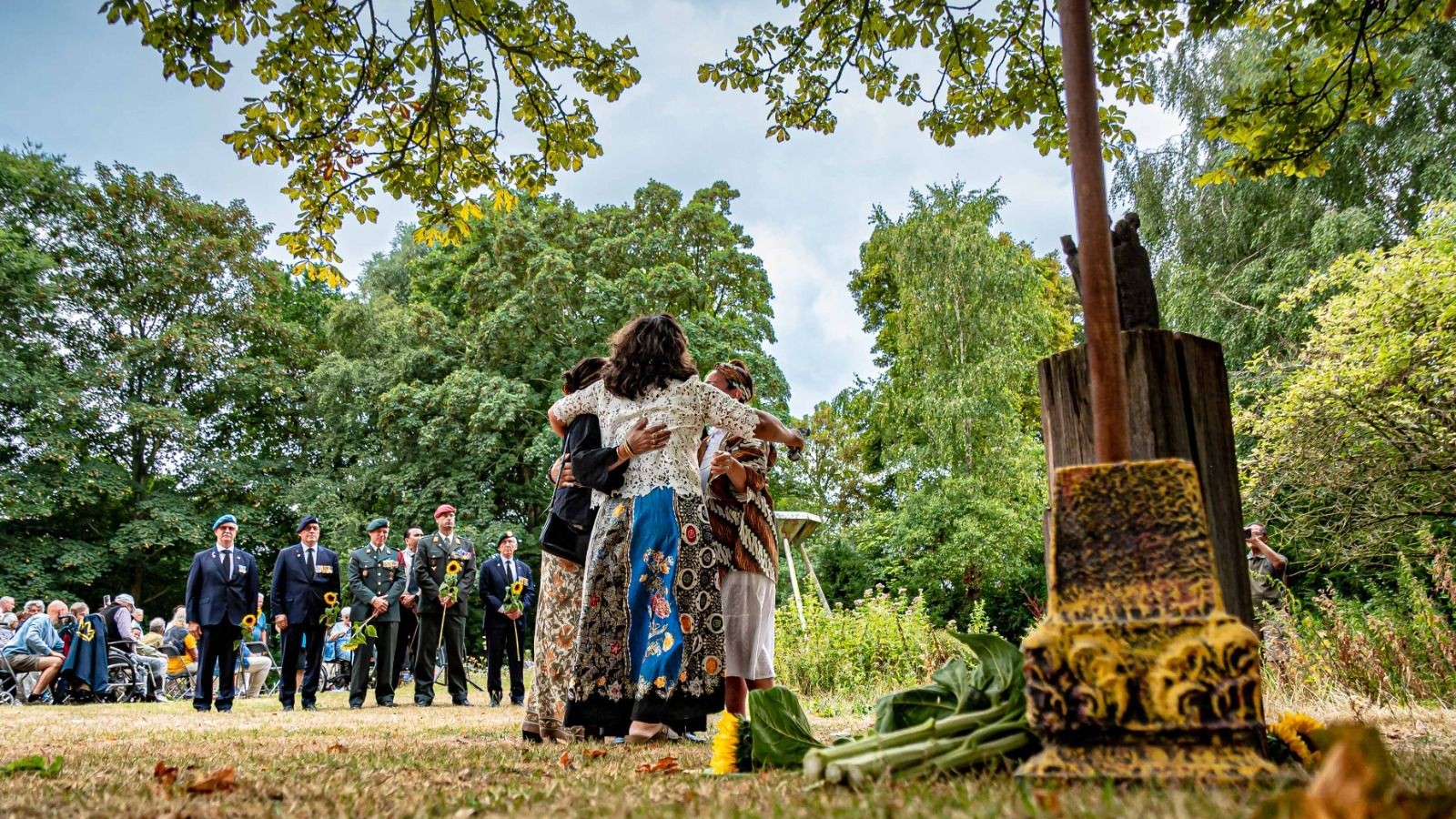 Groep mensen bij houtbewerkt monument in park met bomen, enkele personen in donkere kleding staan naast vrouw in kleurrijke batikkleding,