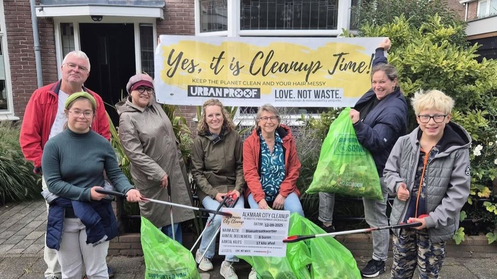 Groep van zes personen staat buiten voor een huis met een banner 'Yes it is Cleanup Time' en houdt opruimgereedschap vast.