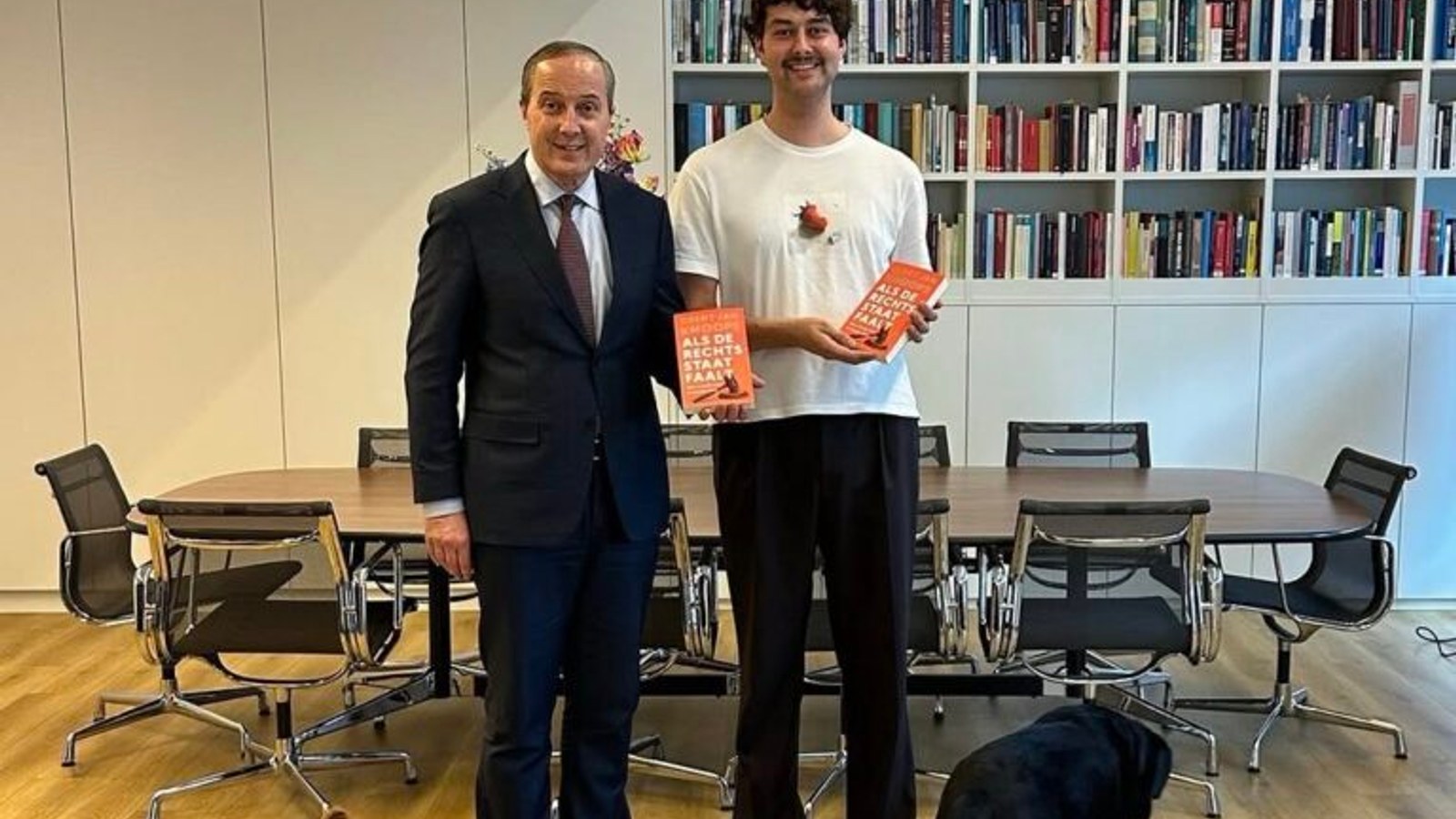 Man in donker pak en man in wit shirt houden rode boeken vast in kantoor met boekenkasten