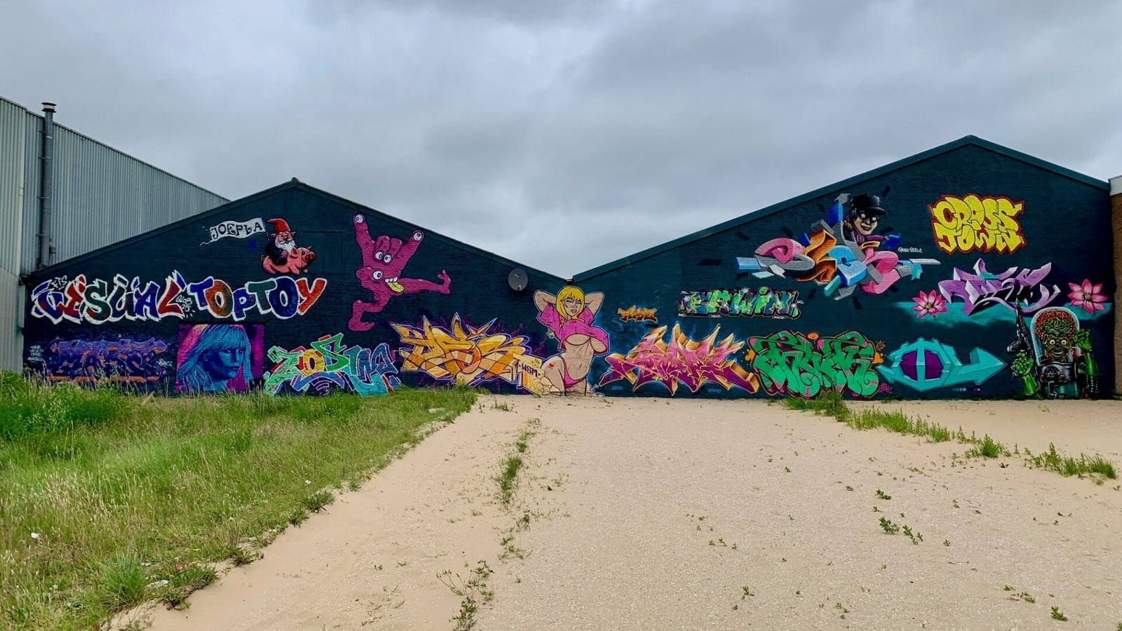 Donkerblauwe gebouwgevel met kleurrijke graffiti en street art schilderingen in roze, geel, groen en paars tegen een bewolkte hemel.