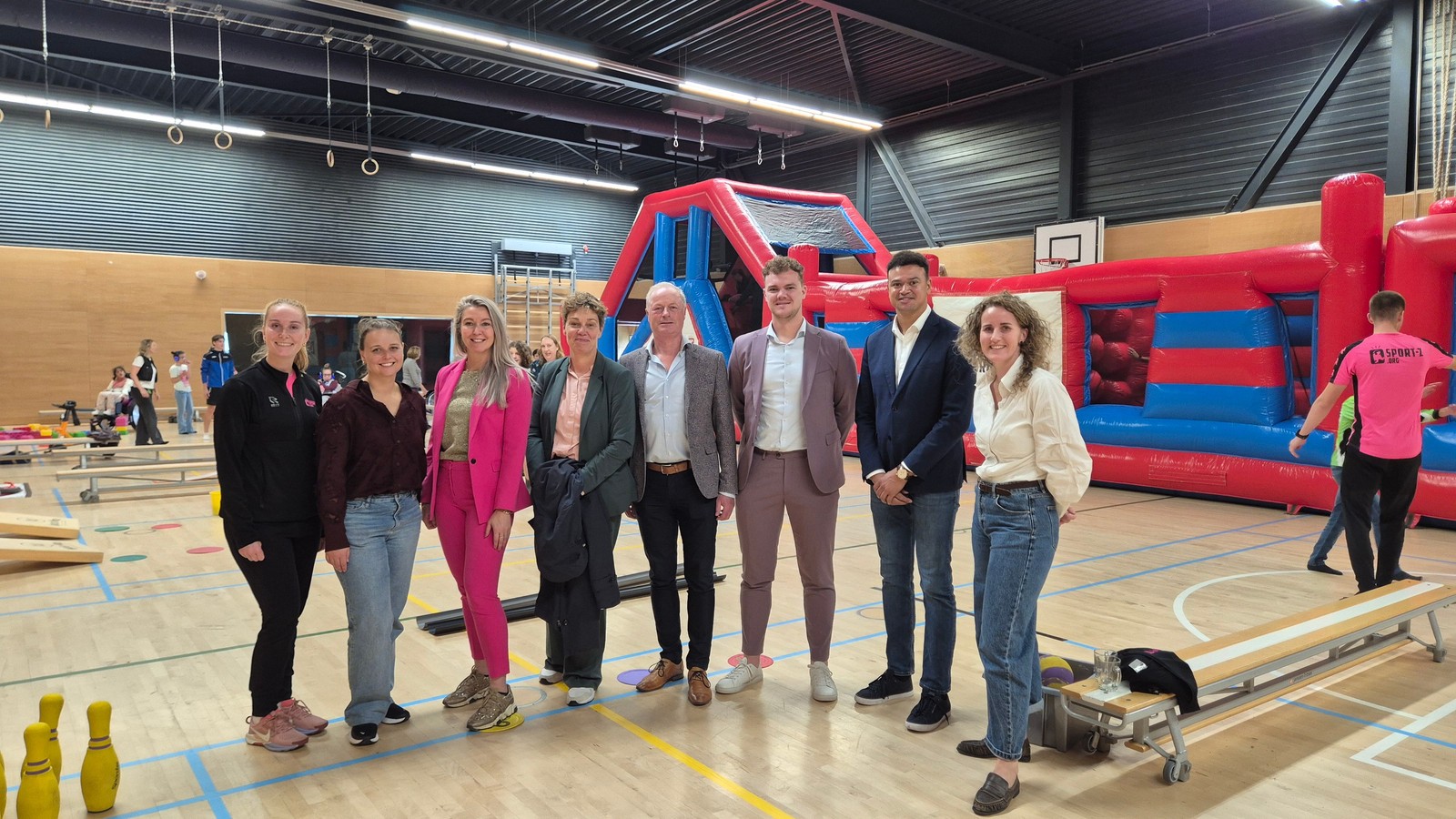 Groep mensen in sporthal met rode opblaasbare speeltoestellen en trampolines op achtergrond