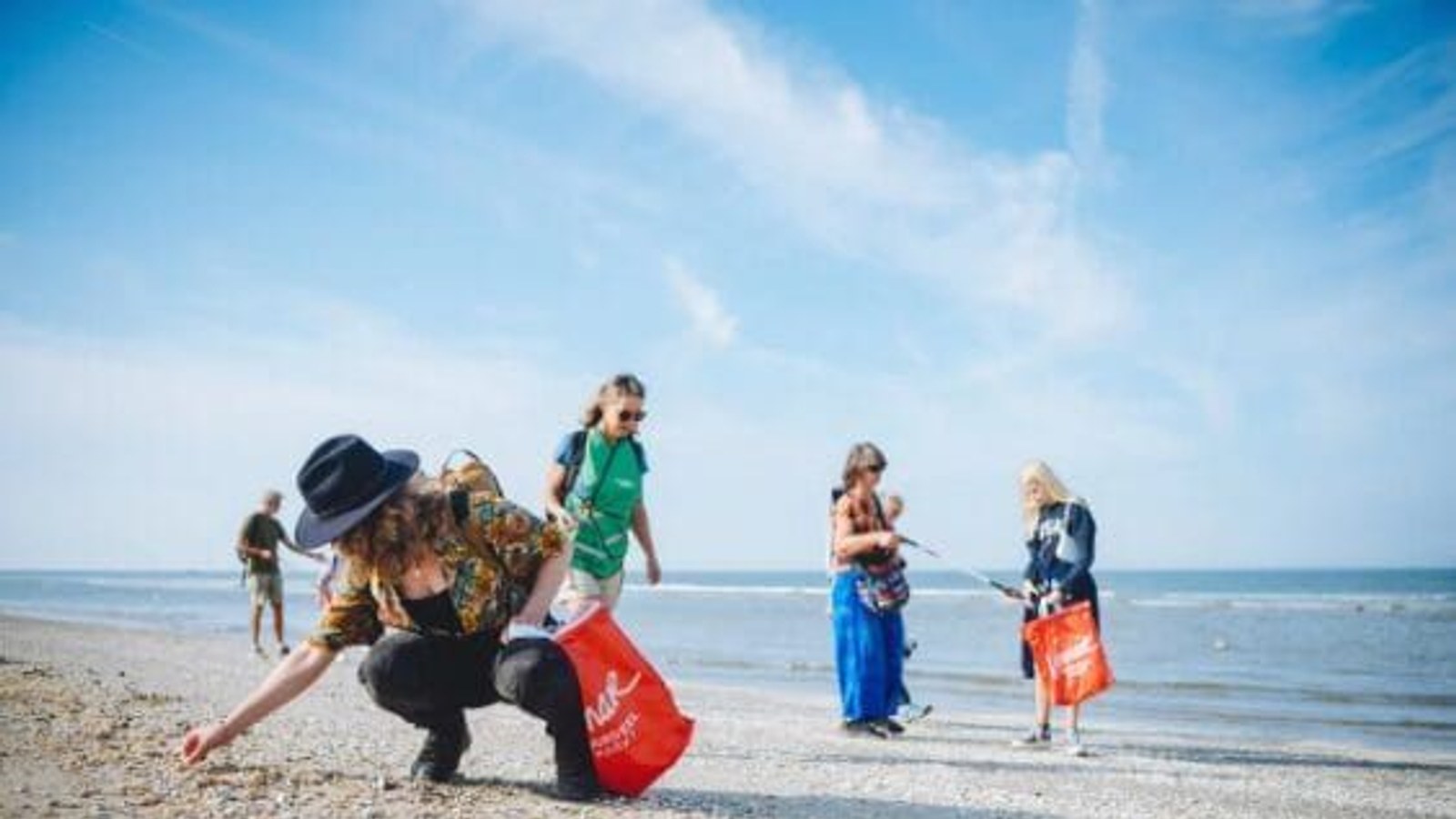 Persoon in groene shirt bukt zich op een zandstrand met bezem en rode emmer om afval op te rapen. Meerdere personen werken mee op de