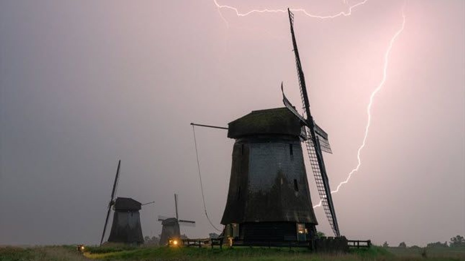 Twee molens in Nederlands landschap onder stormachtige hemel met bliksem, beide molens hebben draaiende wieken