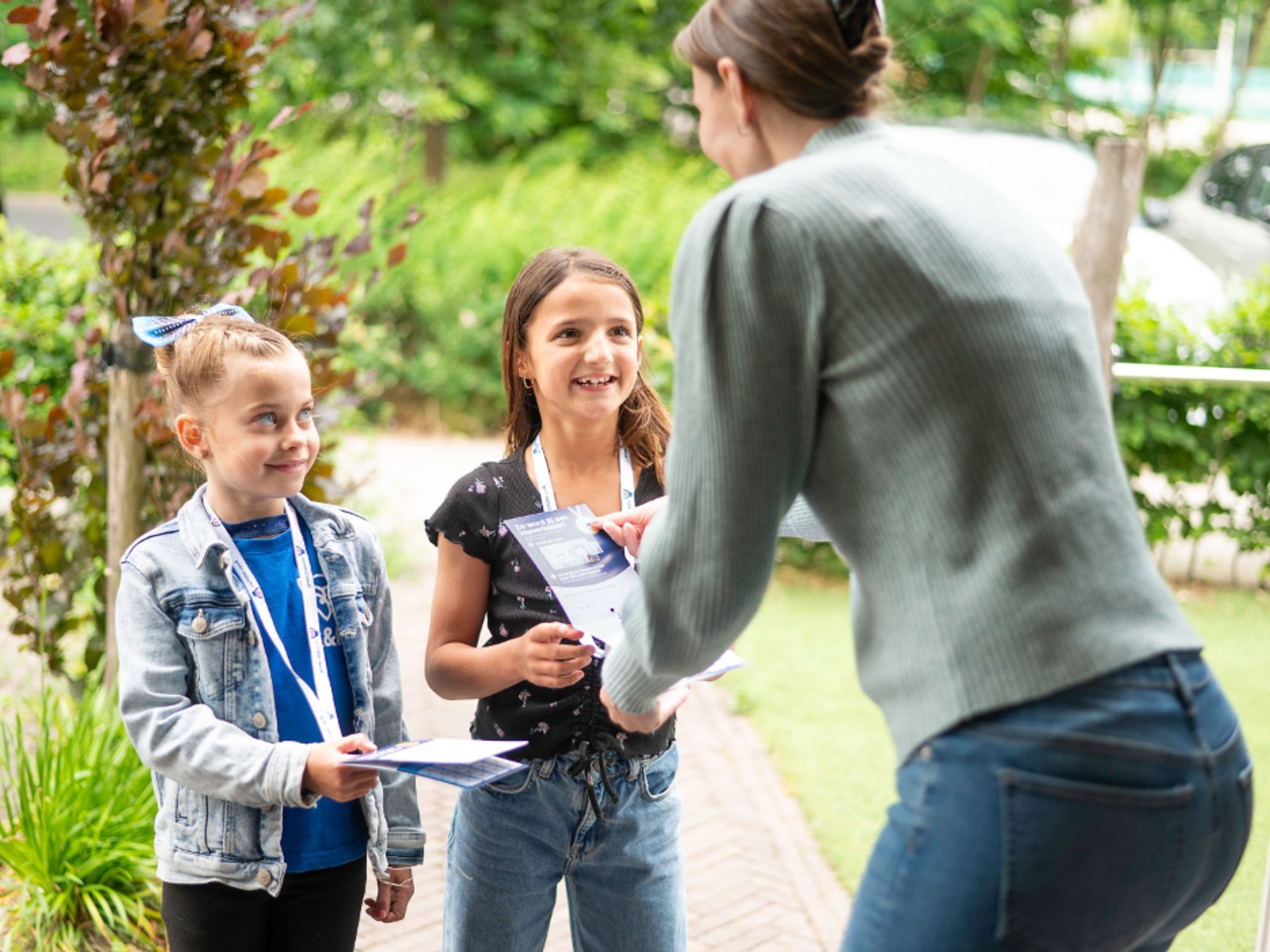 Twee kinderen met halsbanden geven lotjes aan een volwassene in een tuin, voor huizen en groen